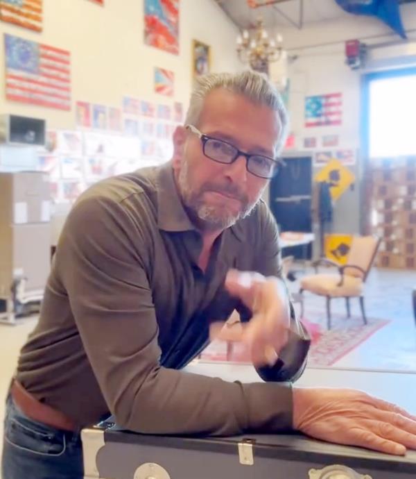 Scott LoBaido in his studio leaning on a table, with Trump traffic signs visible in the background